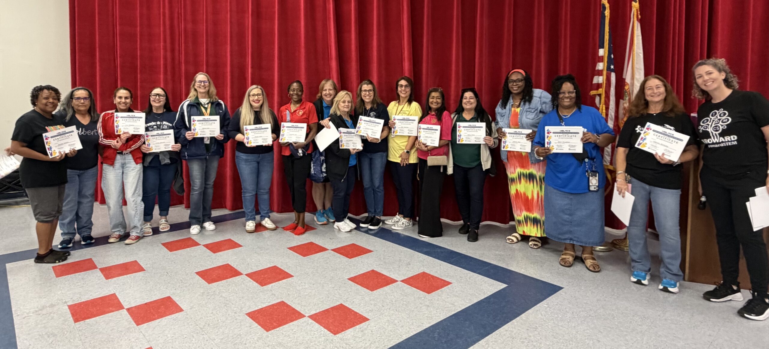 A group of seventeen people standing in a row, most holding certificates, in front of red curtains.